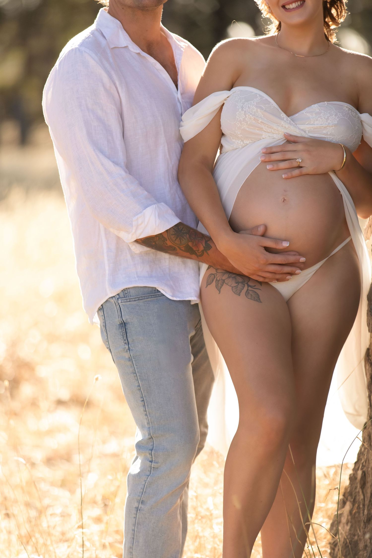 Couple posing outdoors, pregnant woman in white wrap and man in white shirt holding her baby bump in golden field.