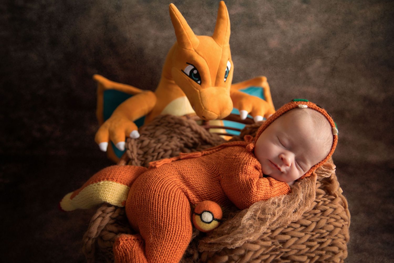 Newborn baby in orange knit Charizard Pokemon onesie sleeping in basket beside Charizard plush toy.