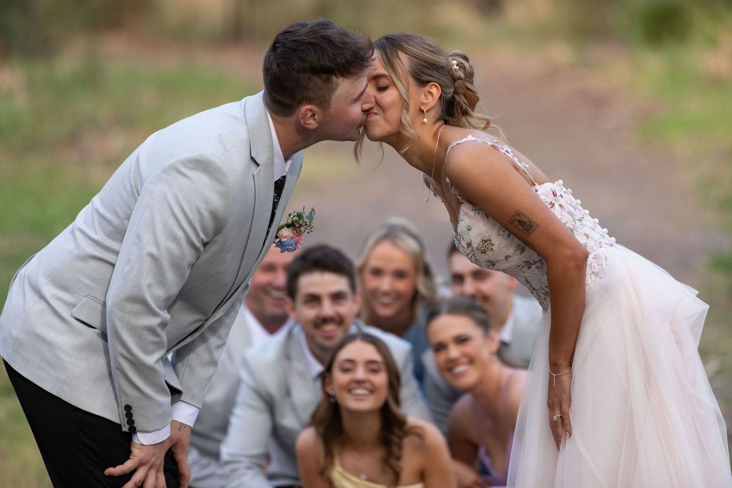 Bride and groom share a romantic kiss surrounded by smiling wedding party guests outdoors.