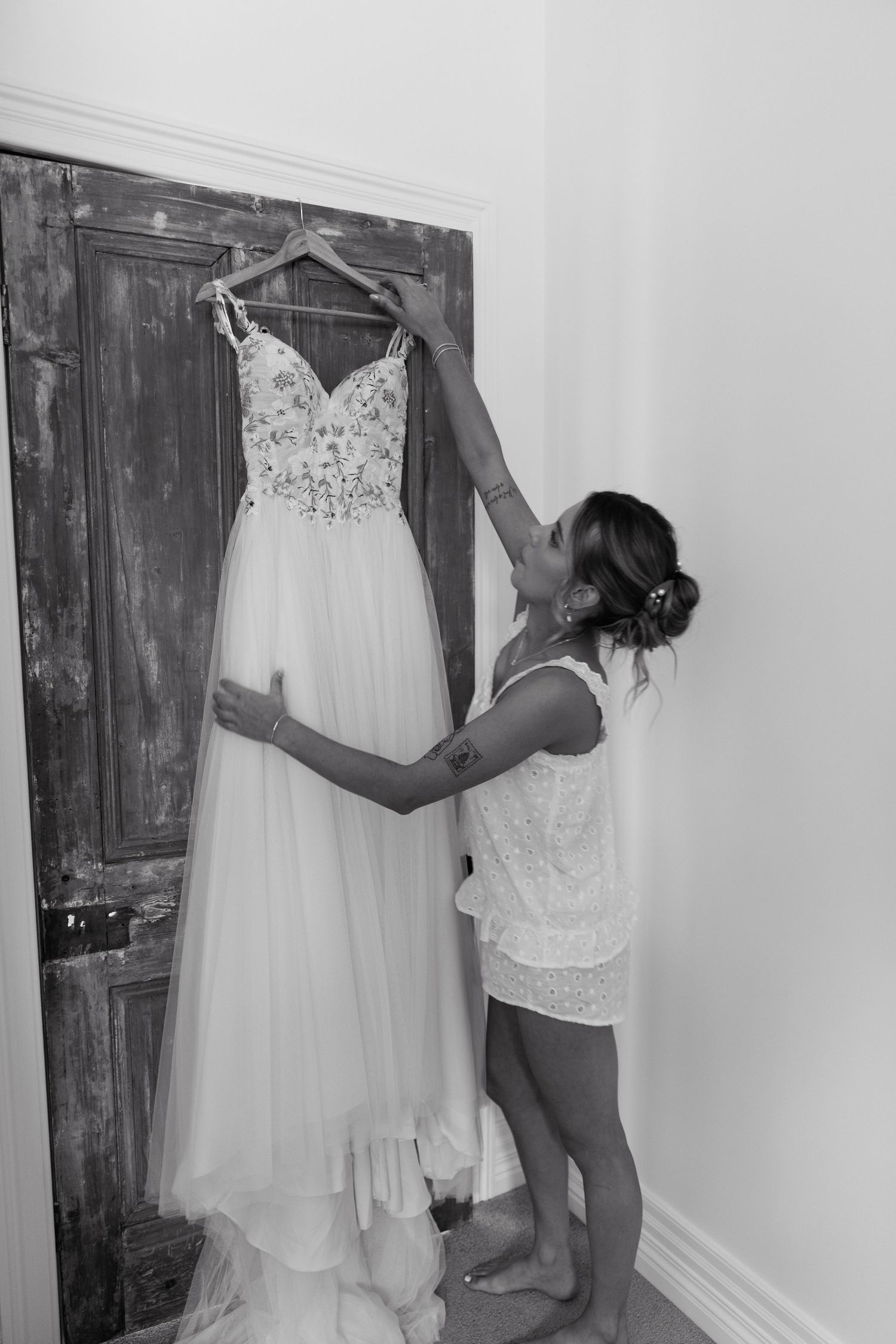 Black and white photo of a bride holding up her elegant lace wedding dress on a hanger against a rustic door.