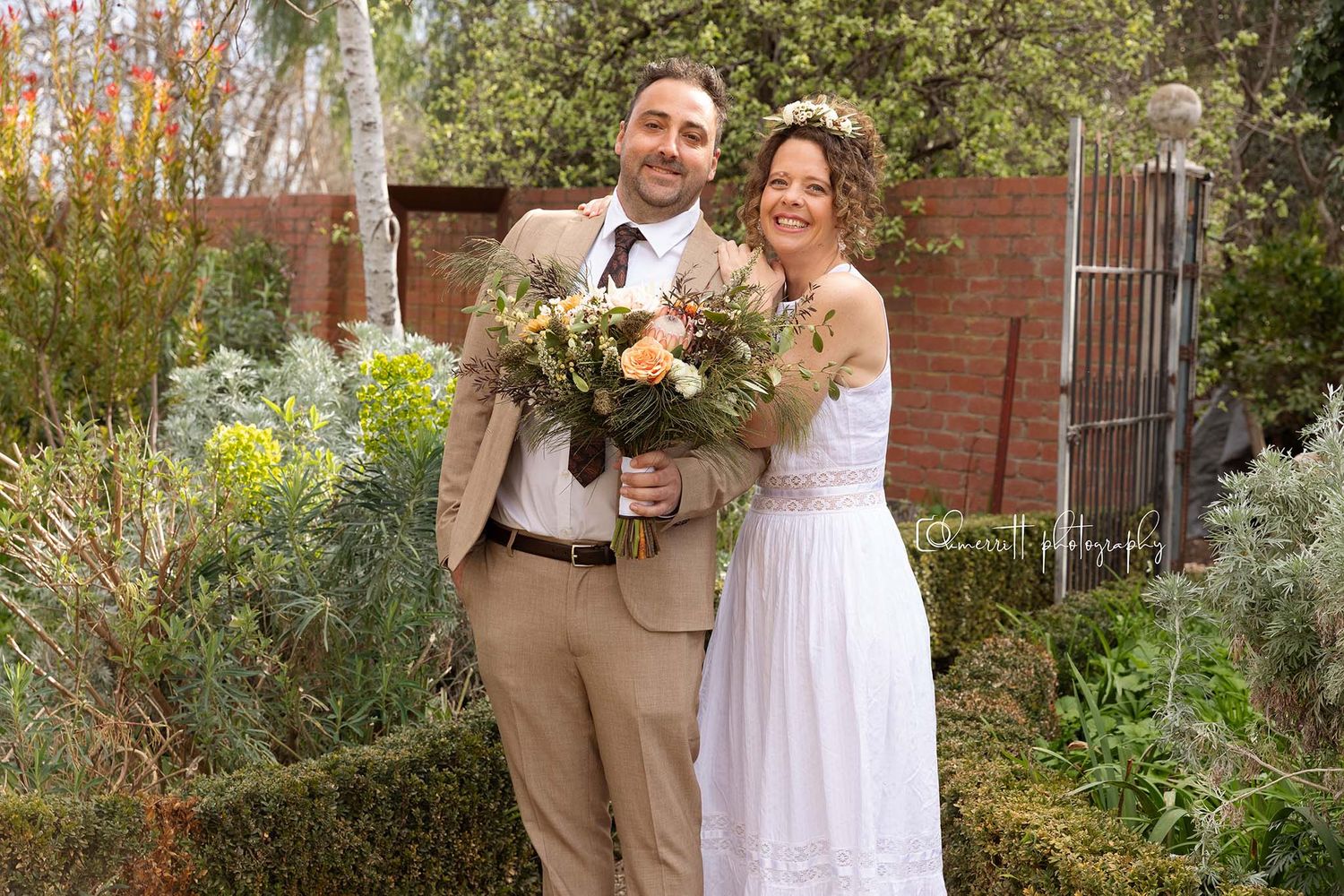Happy couple in garden, groom in tan suit, bride in white dress holding bouquet of orange flowers on their wedding day.
