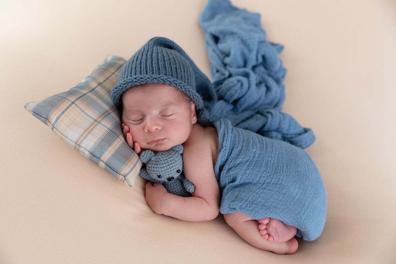Sleeping newborn wearing blue knit hat rests on plaid pillow with soft blue fabric wrap.