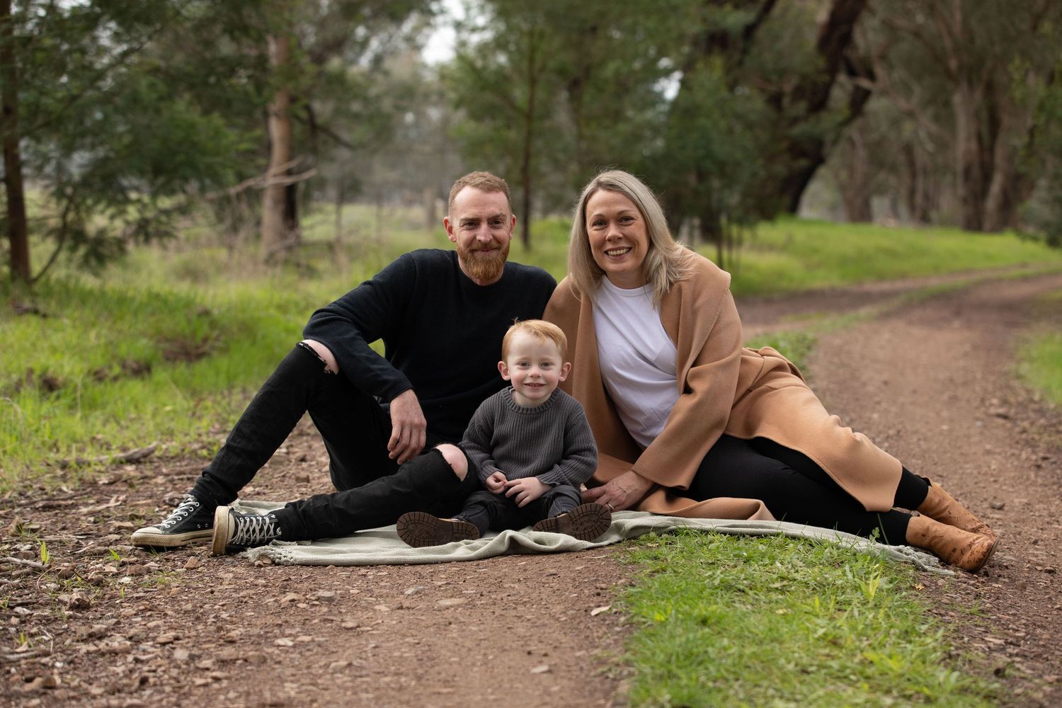 Family of three sitting on a blanket outdoors on a dirt path surrounded by green trees, smiling warmly.