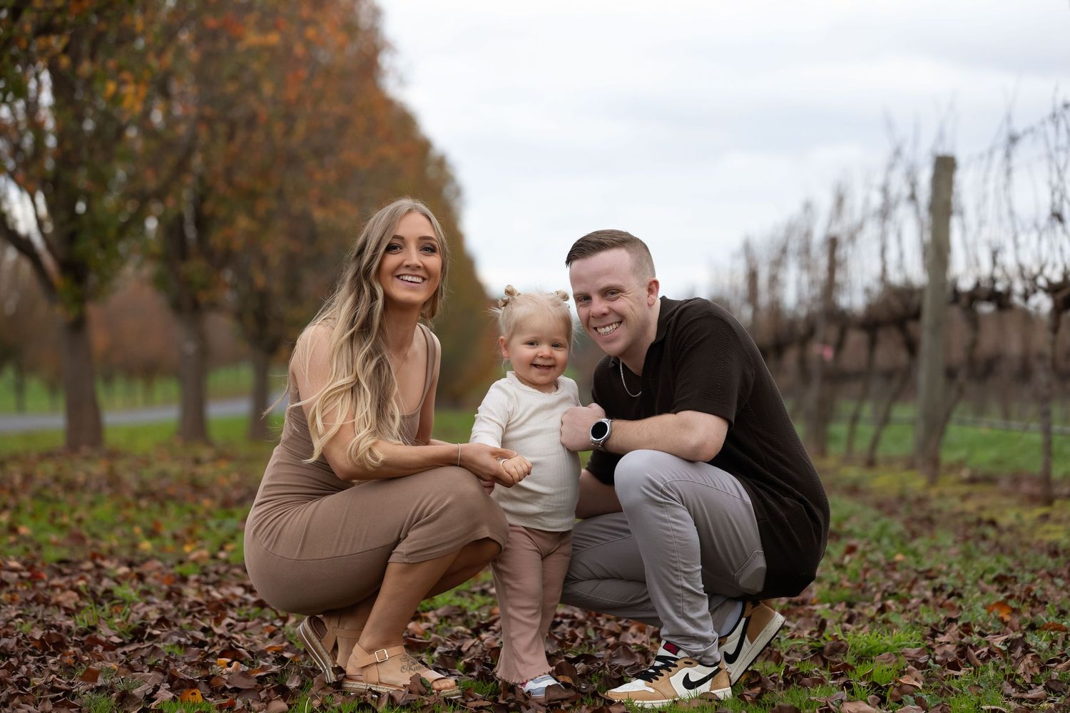 Family portrait of parents with toddler in autumn vineyard setting, surrounded by fallen leaves.