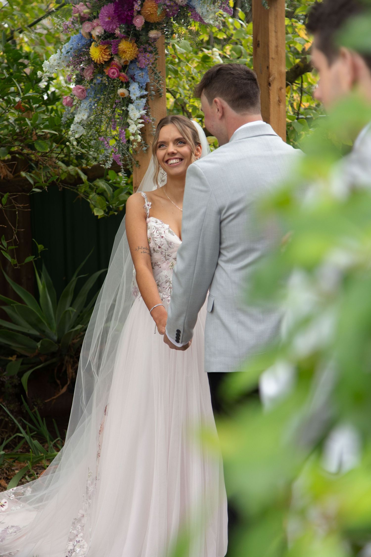 Bride in floral lace gown and veil smiles at groom in grey suit during outdoor garden wedding ceremony.