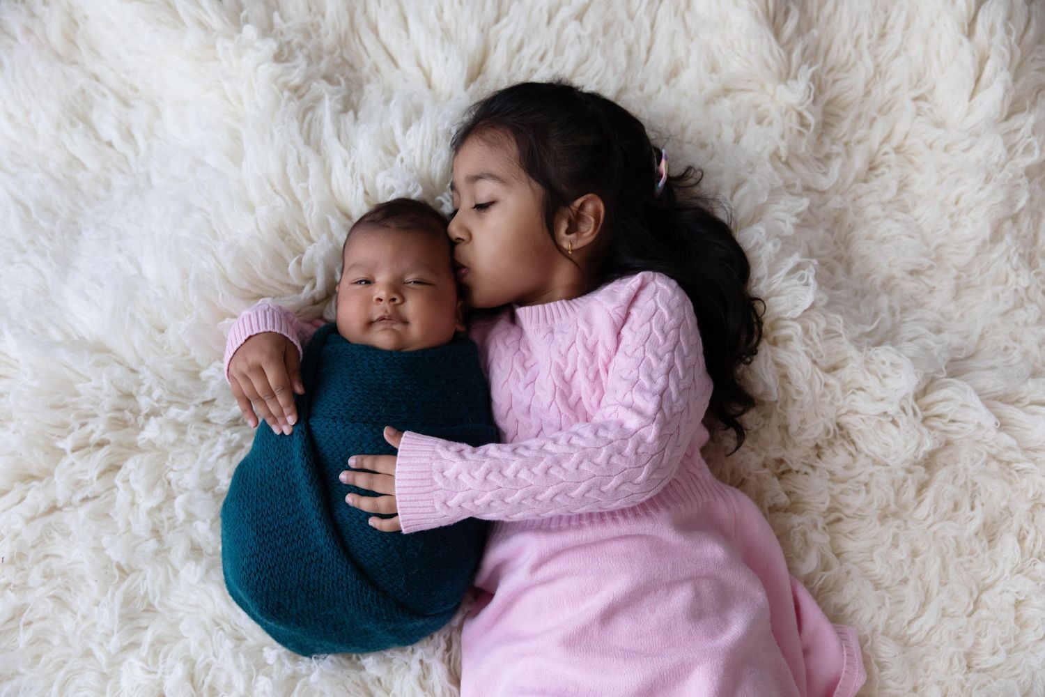 Two children cuddle together on a fluffy white rug, one in a pink sweater and one in a teal outfit.