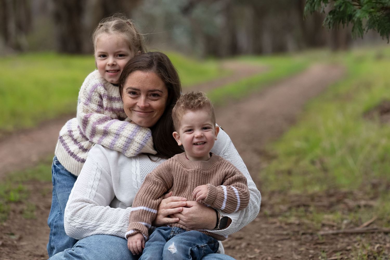 A mother smiles warmly with her two young children on a forest path, all wearing cosy sweaters in a serene outdoor setting.