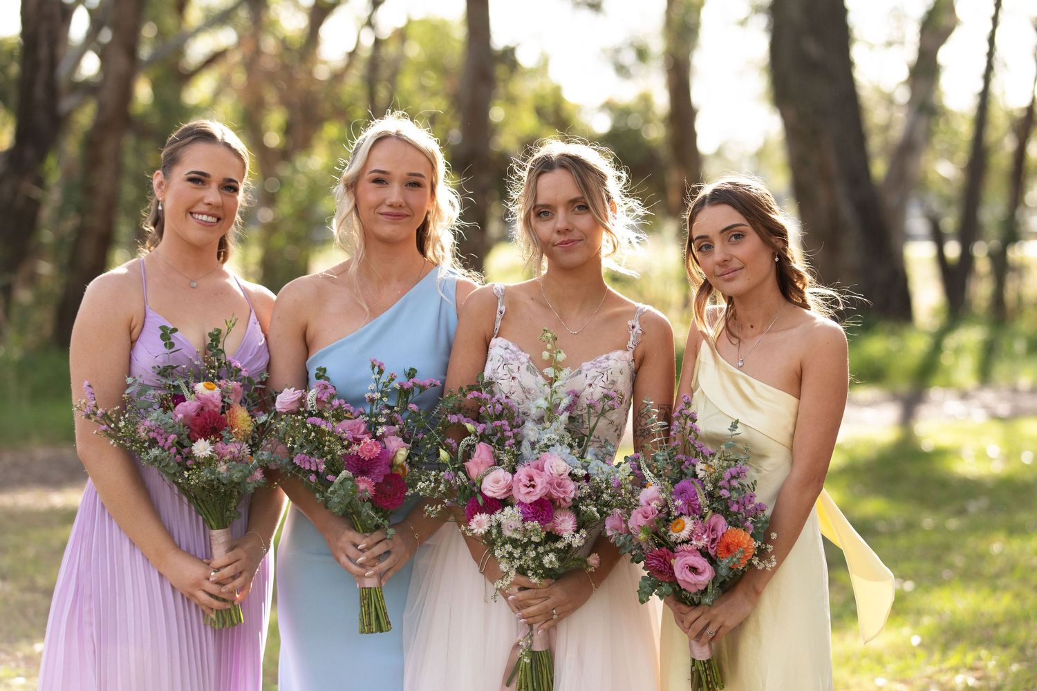 Four bridesmaids in pastel dresses holding colourful floral bouquets, smiling outdoors in a sunlit garden setting.