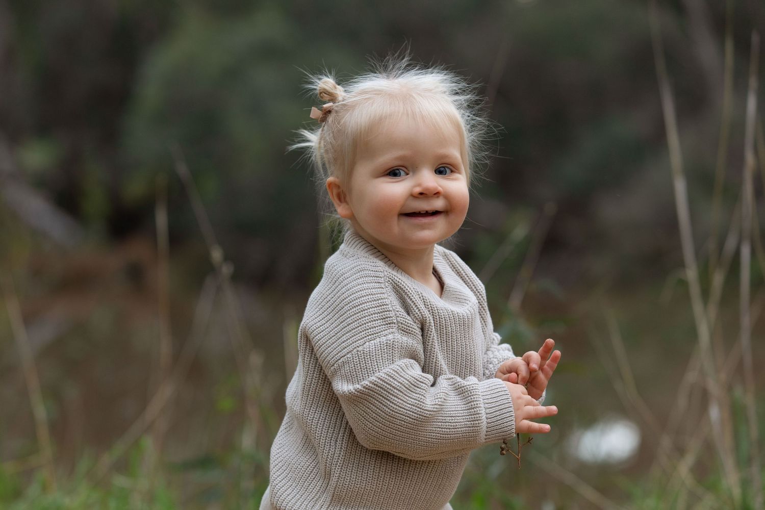 Smiling toddler with wispy blonde hair wearing a beige ribbed sweater, standing outdoors in a natural green setting.