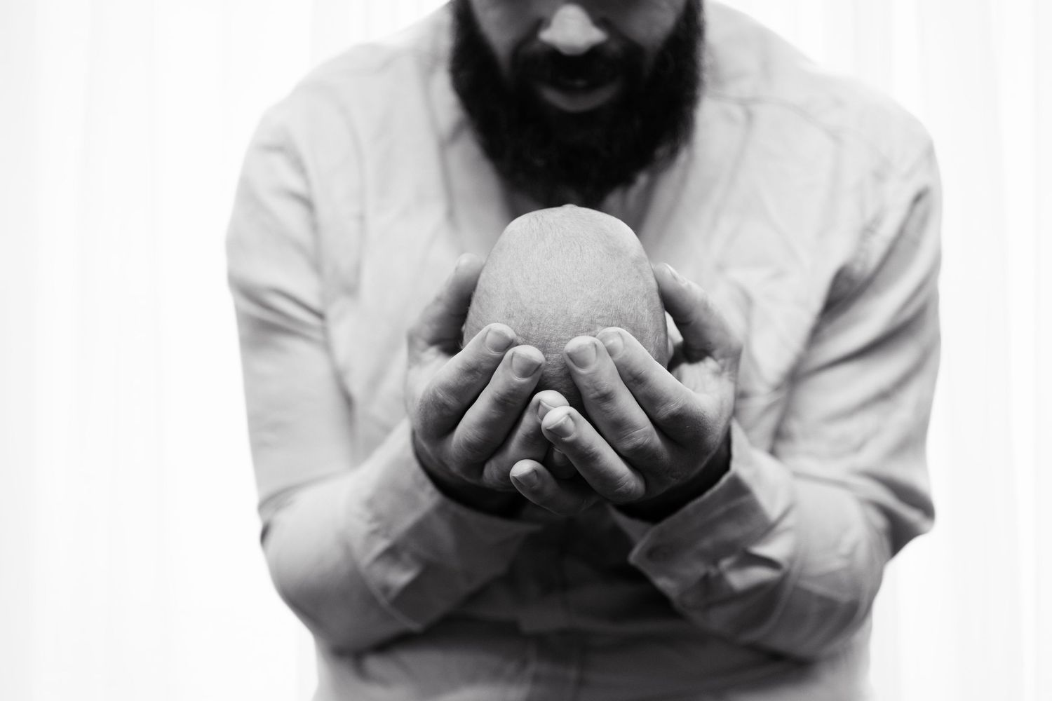 Bearded man in white shirt holding a large egg with both hands in a black and white photo.