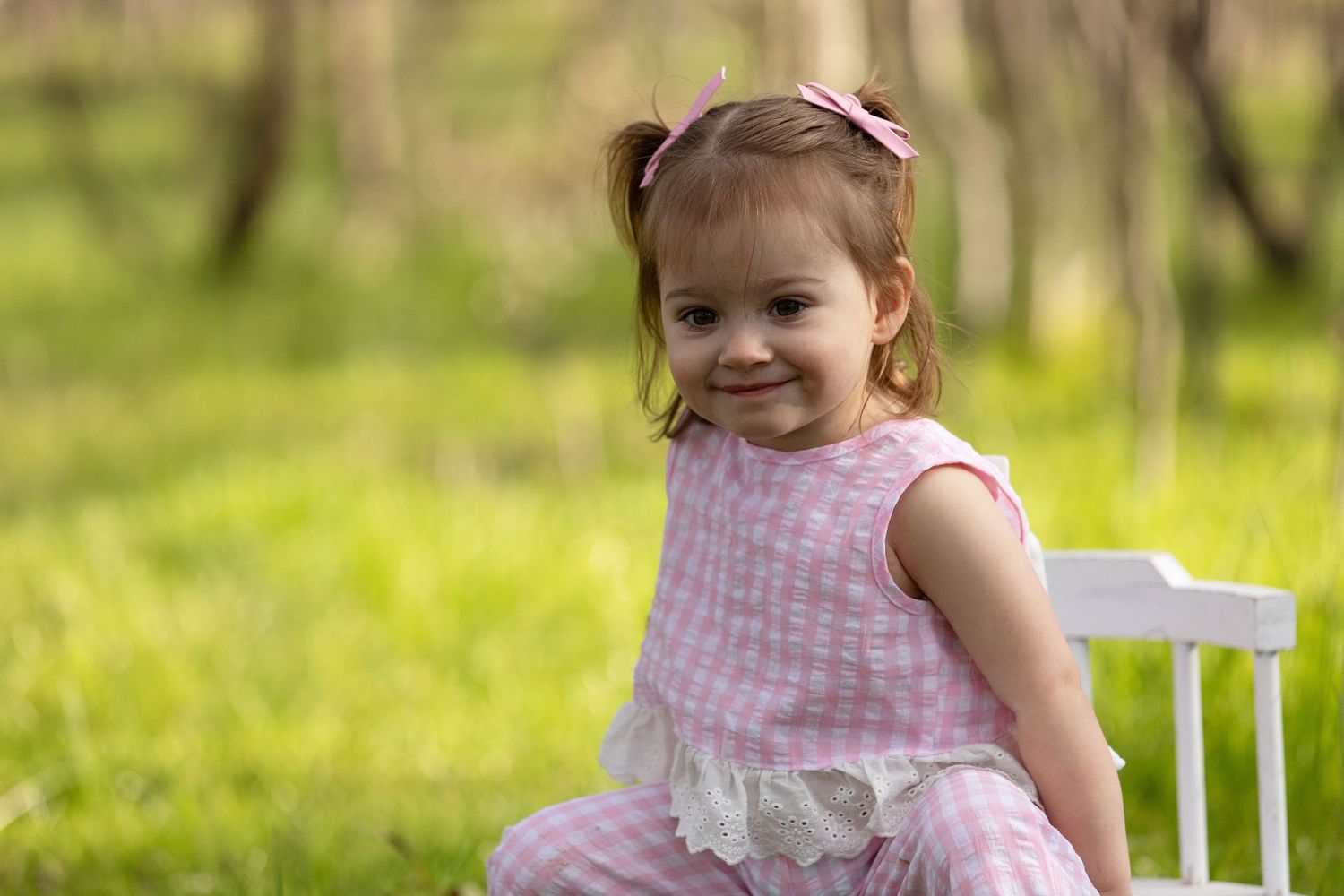 A smiling toddler girl in a pink outfit sits on a white chair outdoors in a lush green park setting.