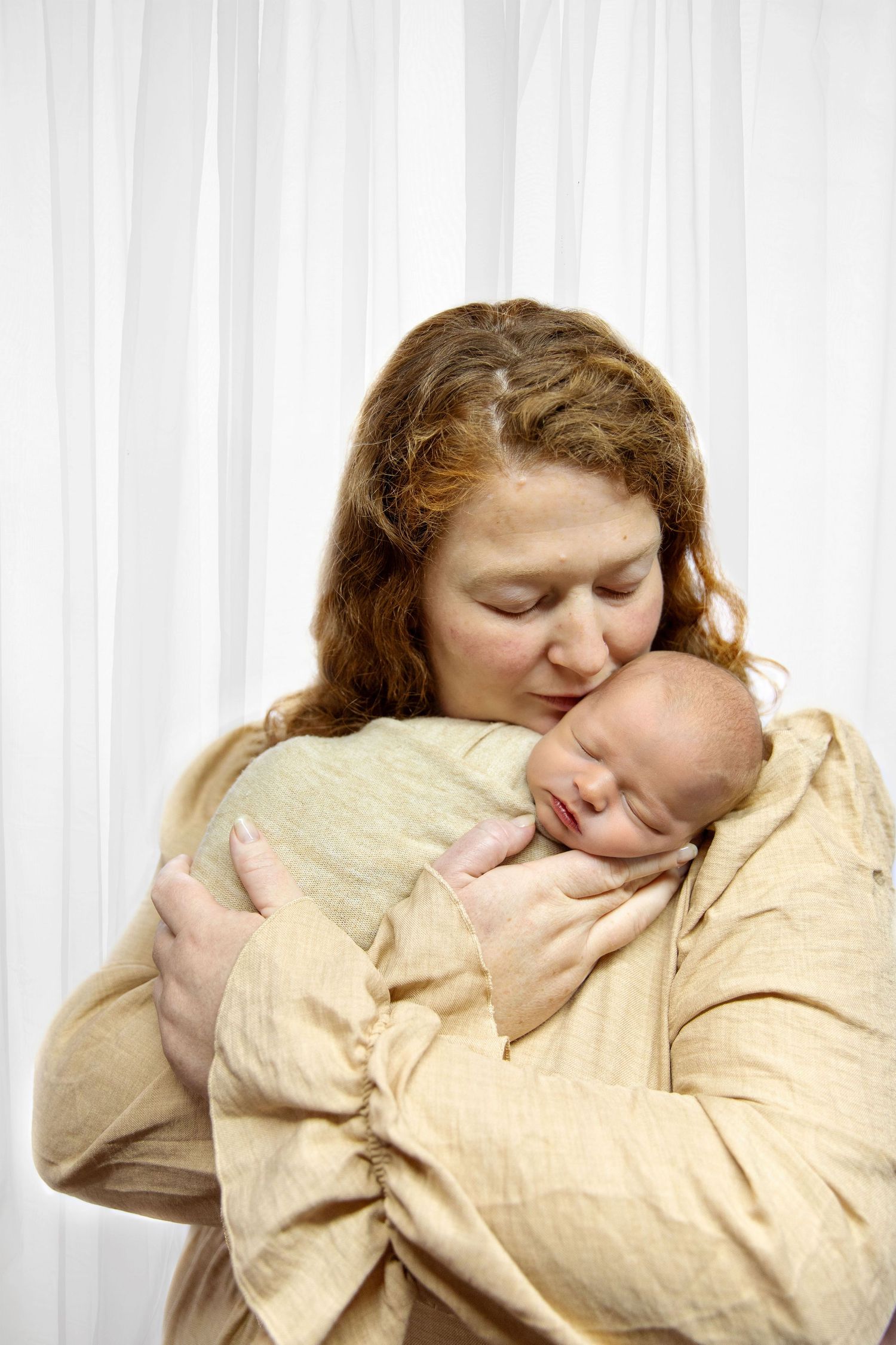 A redheaded mother tenderly kisses her sleeping newborn baby wrapped in a cream blanket against white curtains.