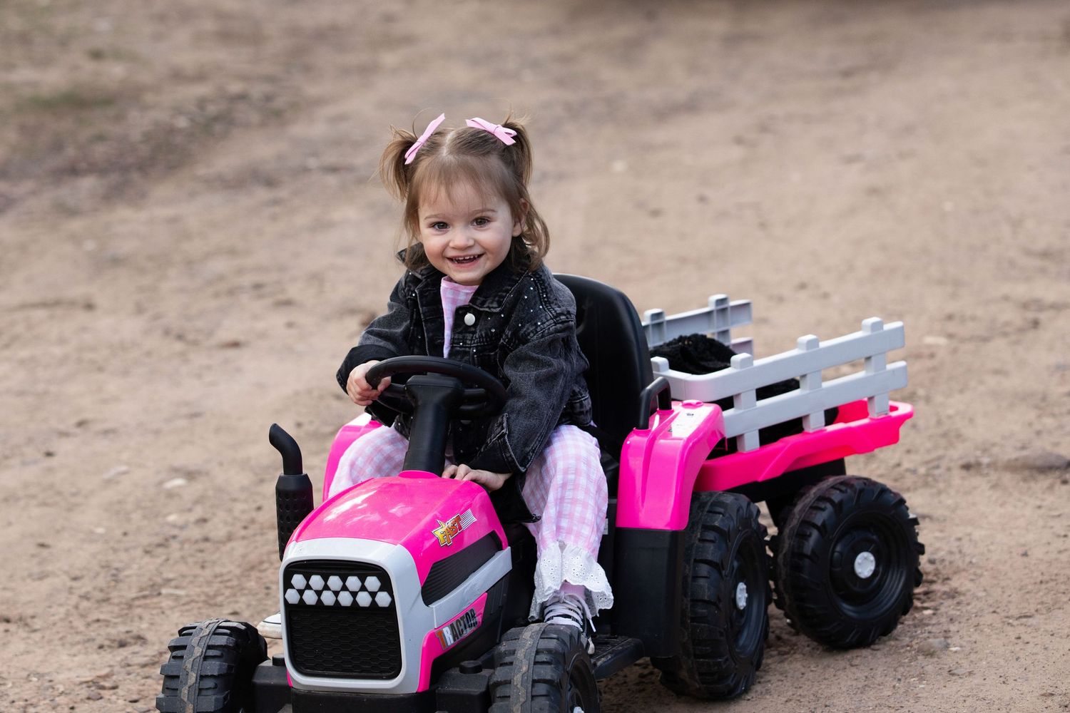 Young girl smiling while riding a pink electric toy tractor with trailer on a dirt path.