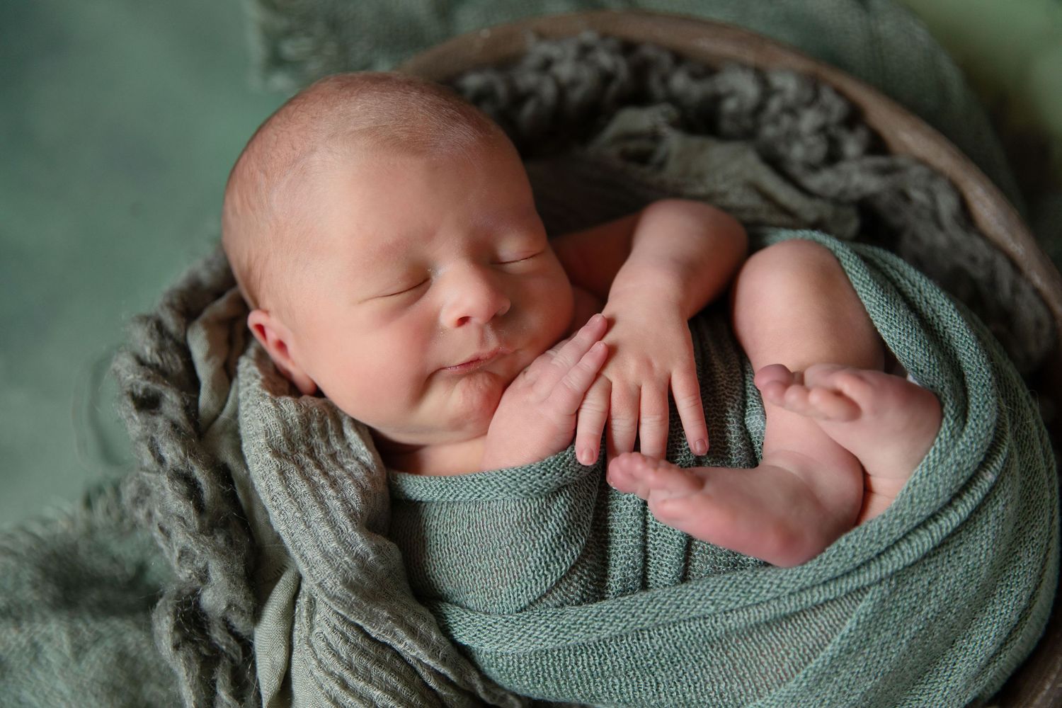 Peaceful newborn baby swaddled in teal wrap sleeping in a wooden bowl with soft green blanket backdrop.