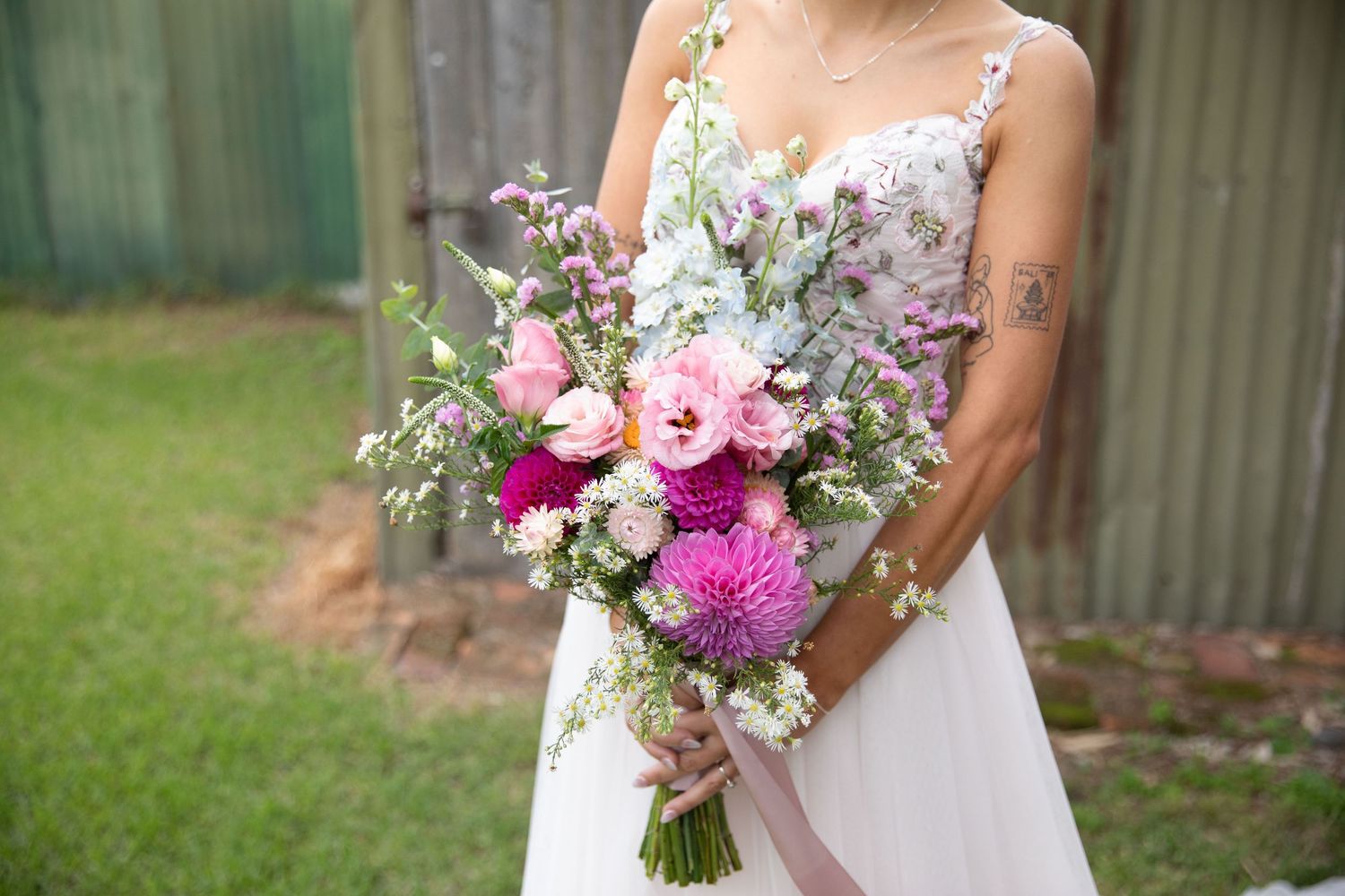 Bride holding a lush pink and purple wedding bouquet with roses and thistles, wearing a white lace gown outdoors.