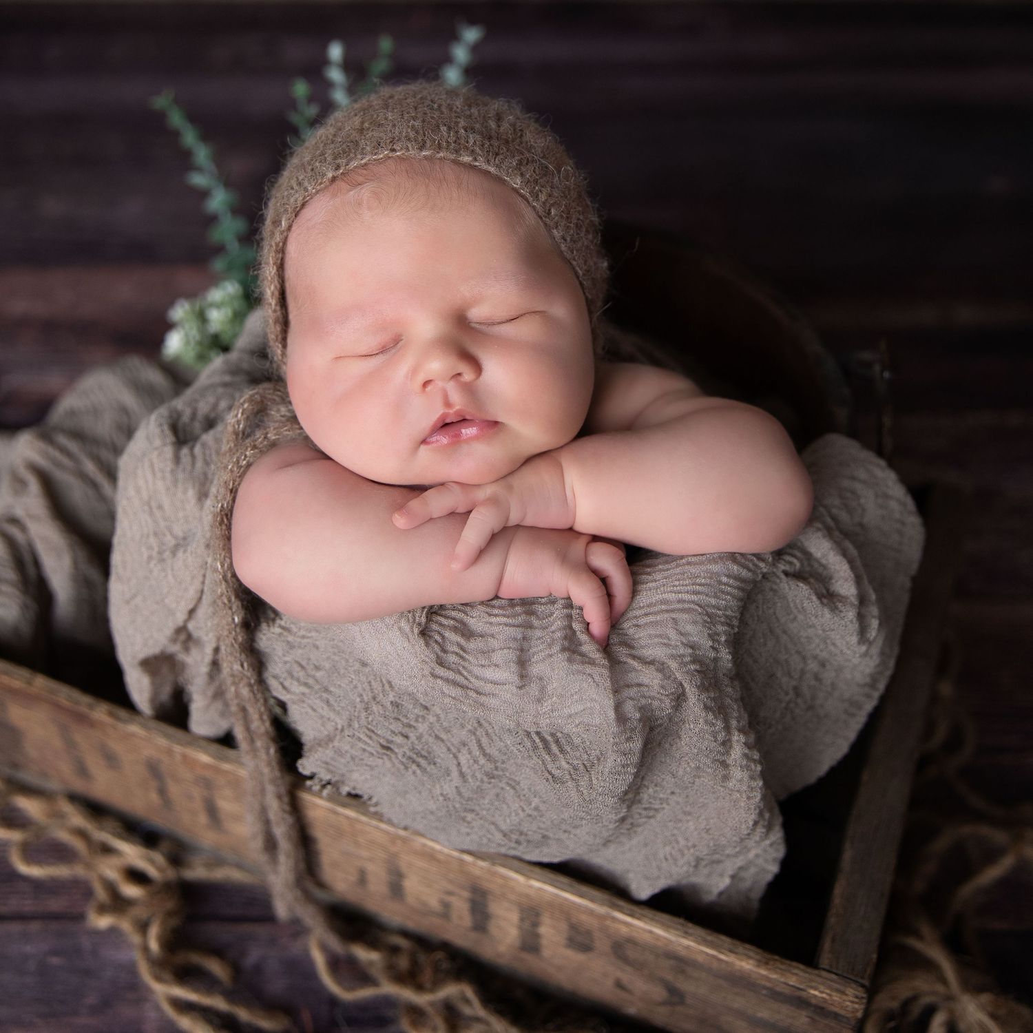Newborn baby sleeping peacefully in a wooden crate, wrapped in a soft taupe blanket and wearing a knit bonnet.