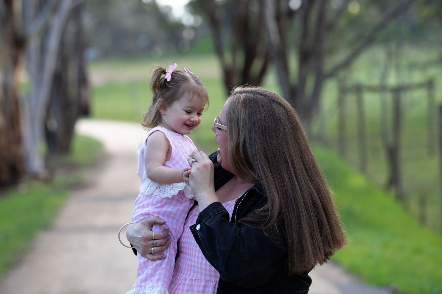 A mother lovingly holds her smiling toddler daughter on a tree-lined path outdoors.