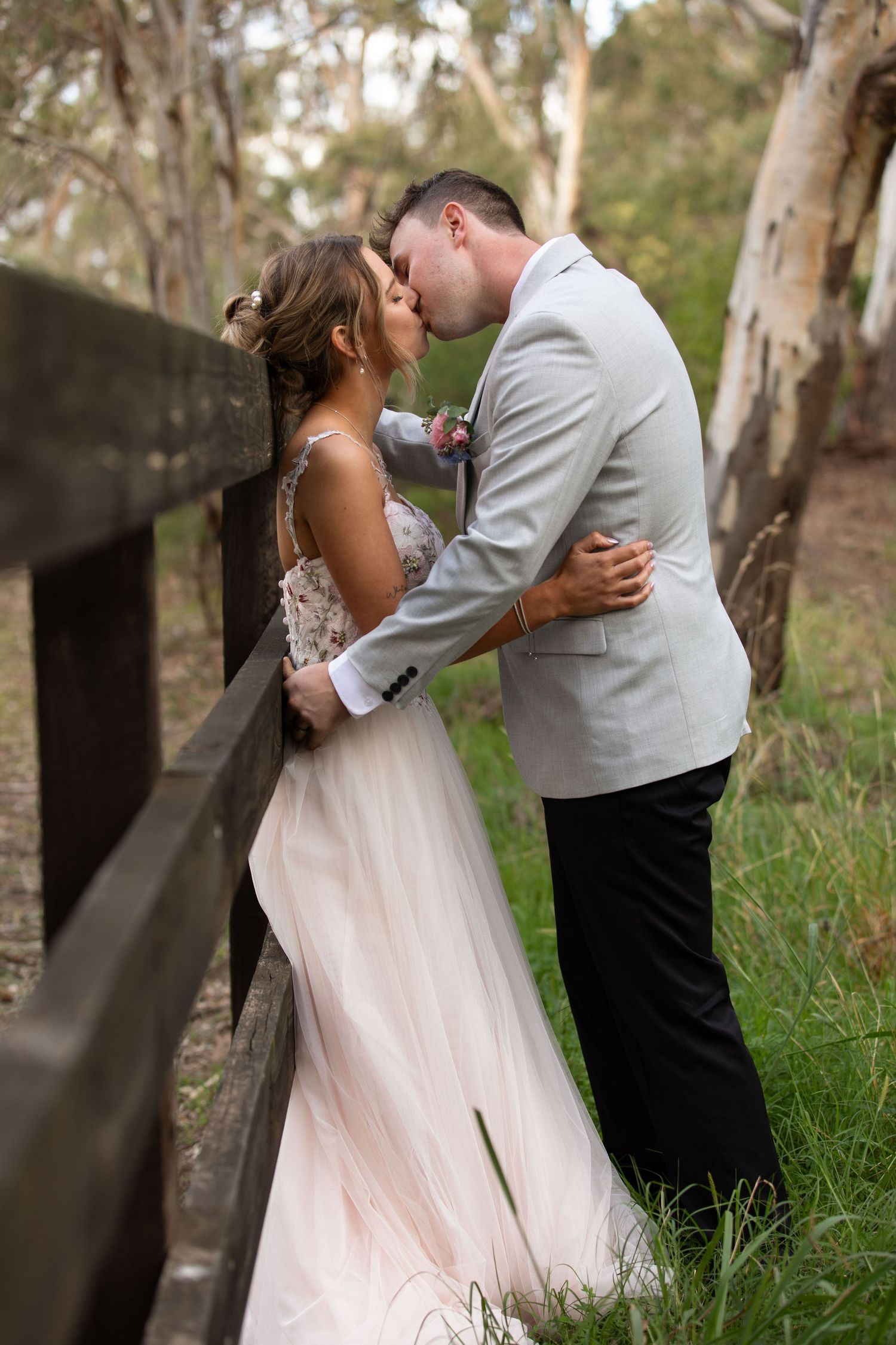 Bride and groom share a romantic kiss beside a wooden fence surrounded by lush greenery in an outdoor wedding setting.