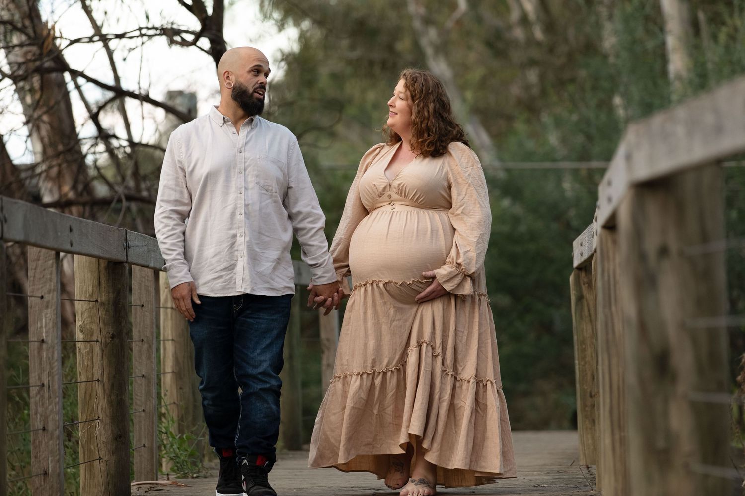 Pregnant couple holding hands walking on a wooden bridge path outdoors during a maternity photo shoot.