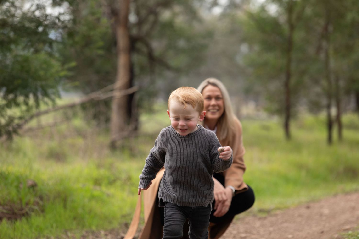 A smiling blonde woman crouches behind a happy toddler boy running on a dirt path in a lush green forest.