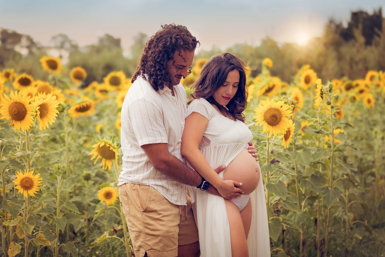 A romantic maternity photo taken in a sunflower field at golden hour shows a gentle embrace against the setting sun.
