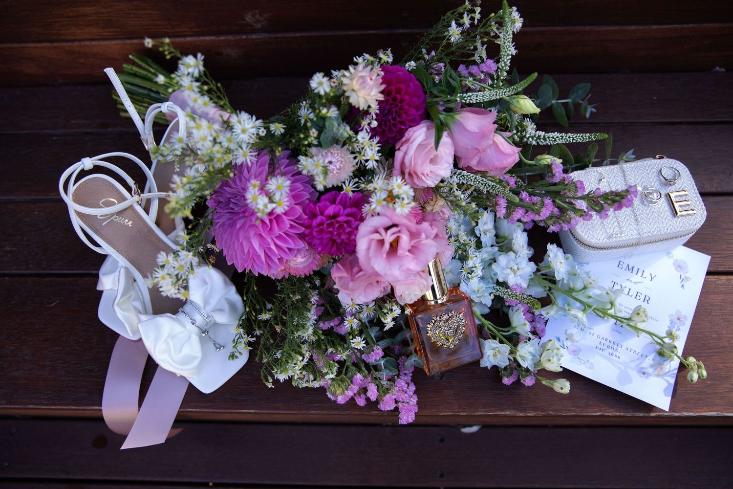 Purple and pink floral wedding bouquet with sandals and invitation on rustic wooden surface.