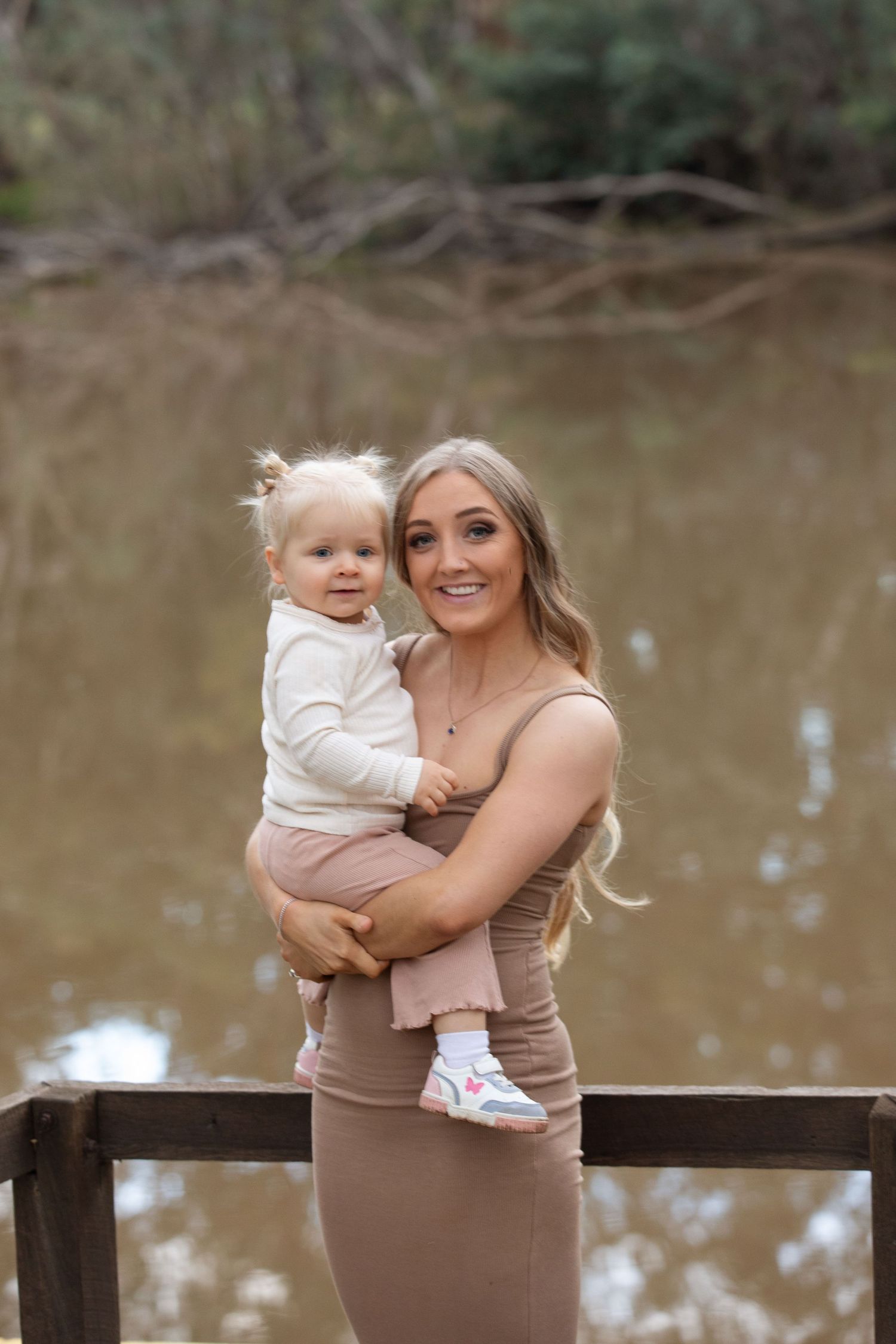 A smiling young woman holds a toddler on a wooden dock with a calm pond and bare trees in the background.