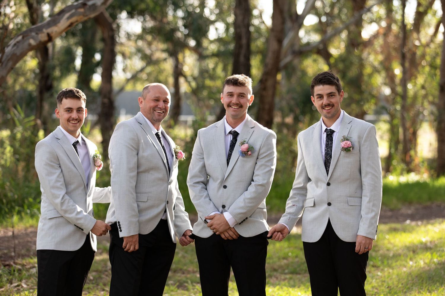 Four groomsmen in white blazers and dark ties smiling outdoors in a lush green forest setting.