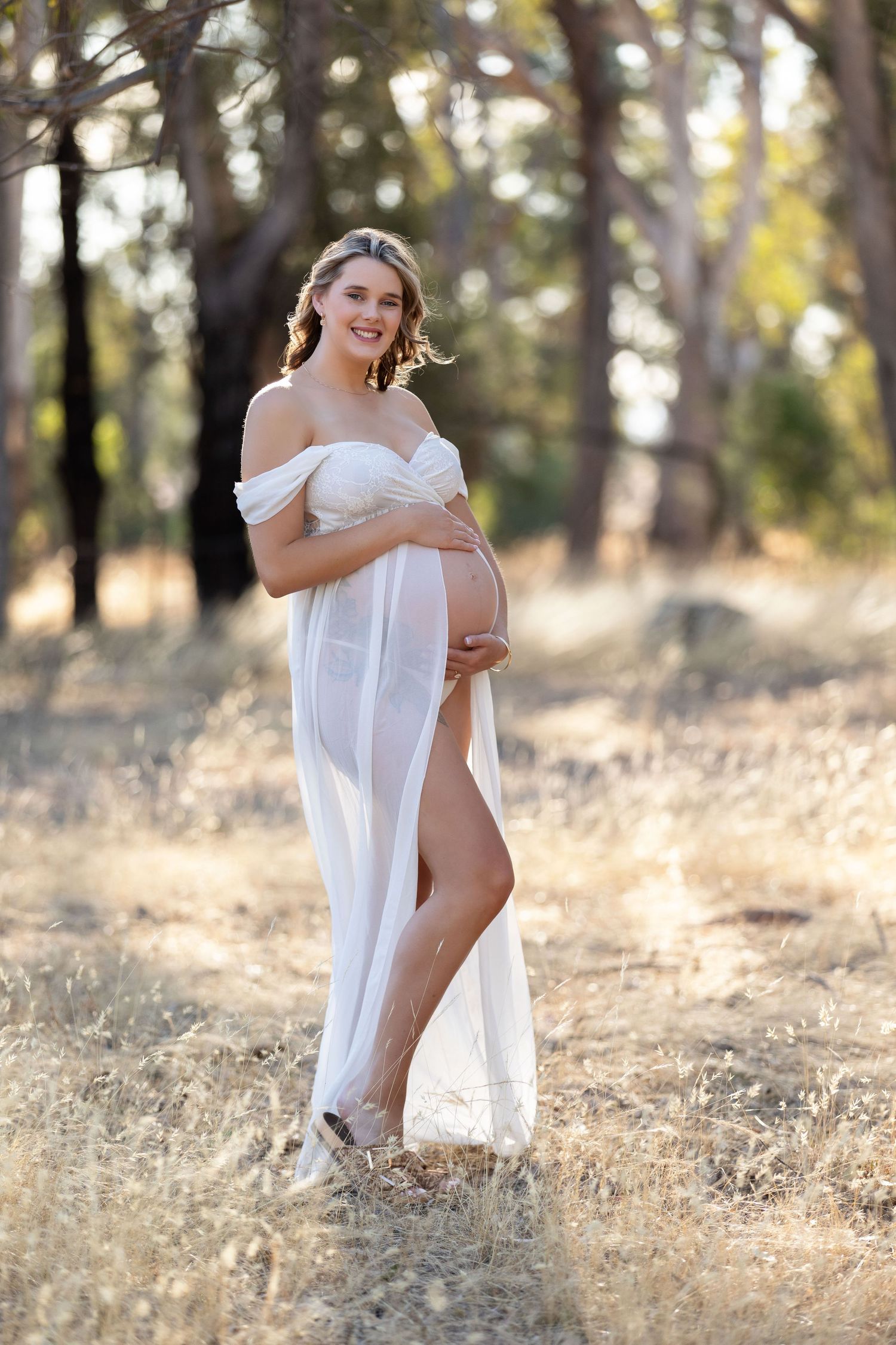 Smiling pregnant woman in flowing white off-shoulder gown cradling her belly outdoors in a sunlit forest setting.