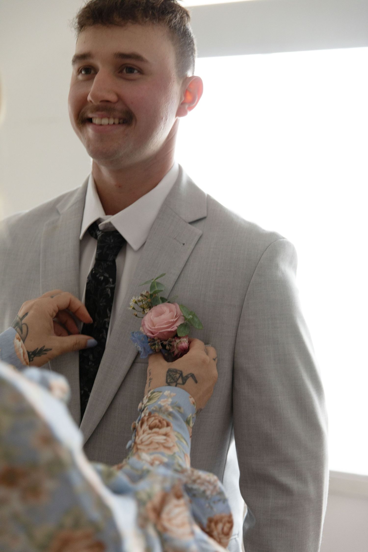 Smiling groom in grey suit with floral boutonniere being pinned on lapel by tattooed hands at wedding.