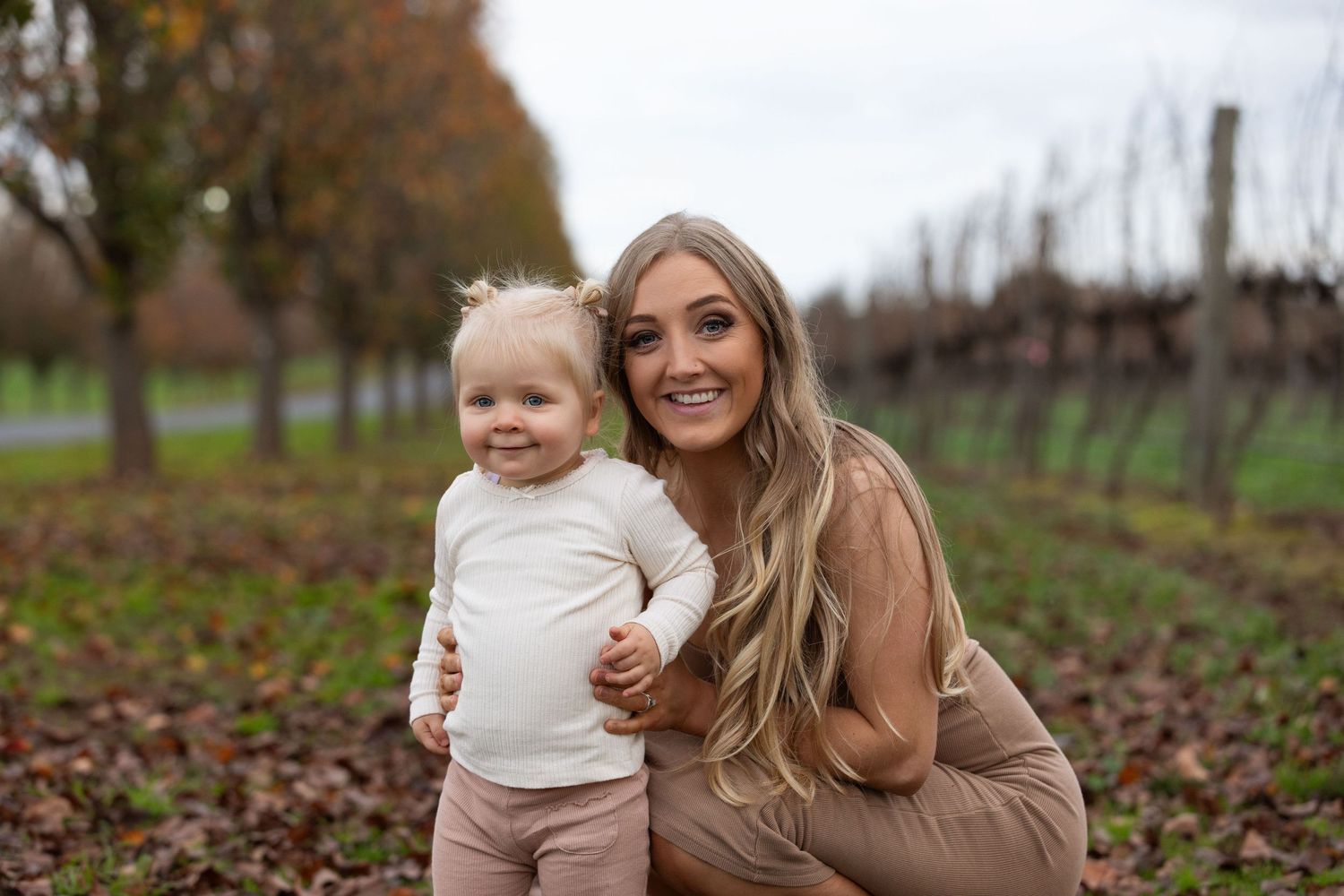 A young woman crouches beside a smiling toddler in a vineyard during autumn, both dressed in neutral tones.