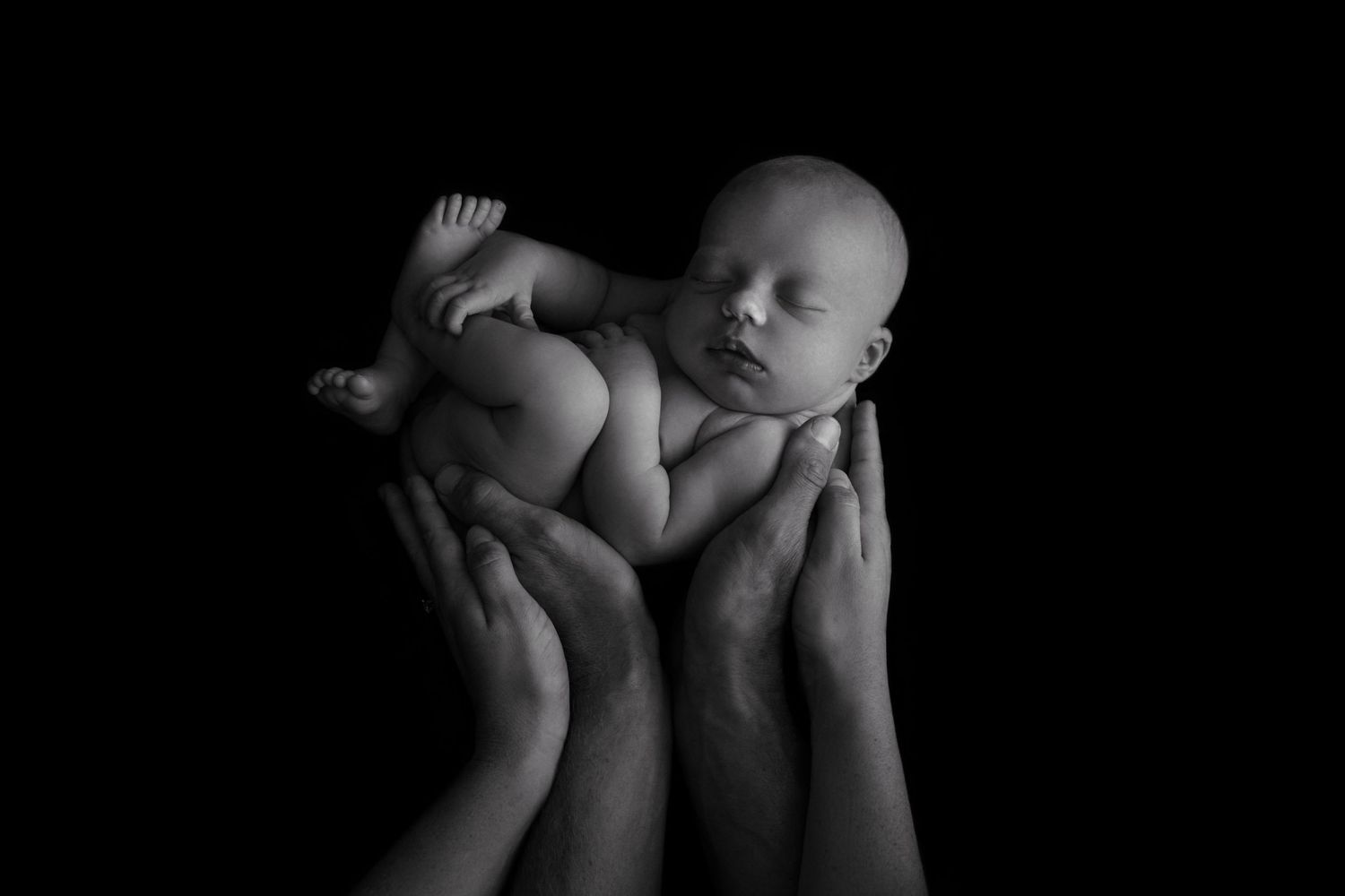 Adult hands tenderly hold a sleeping newborn baby against a dark background in dramatic black and white.