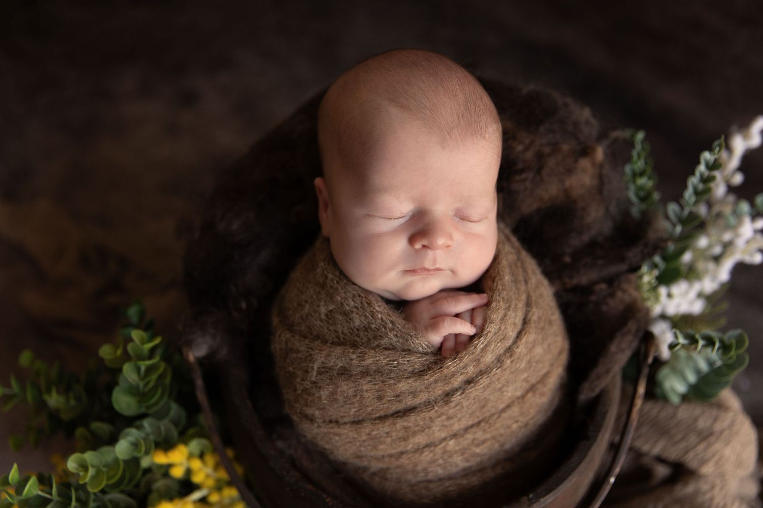 Sleeping newborn baby wrapped in brown knit swaddle inside rustic wooden bucket with green and yellow floral accents.