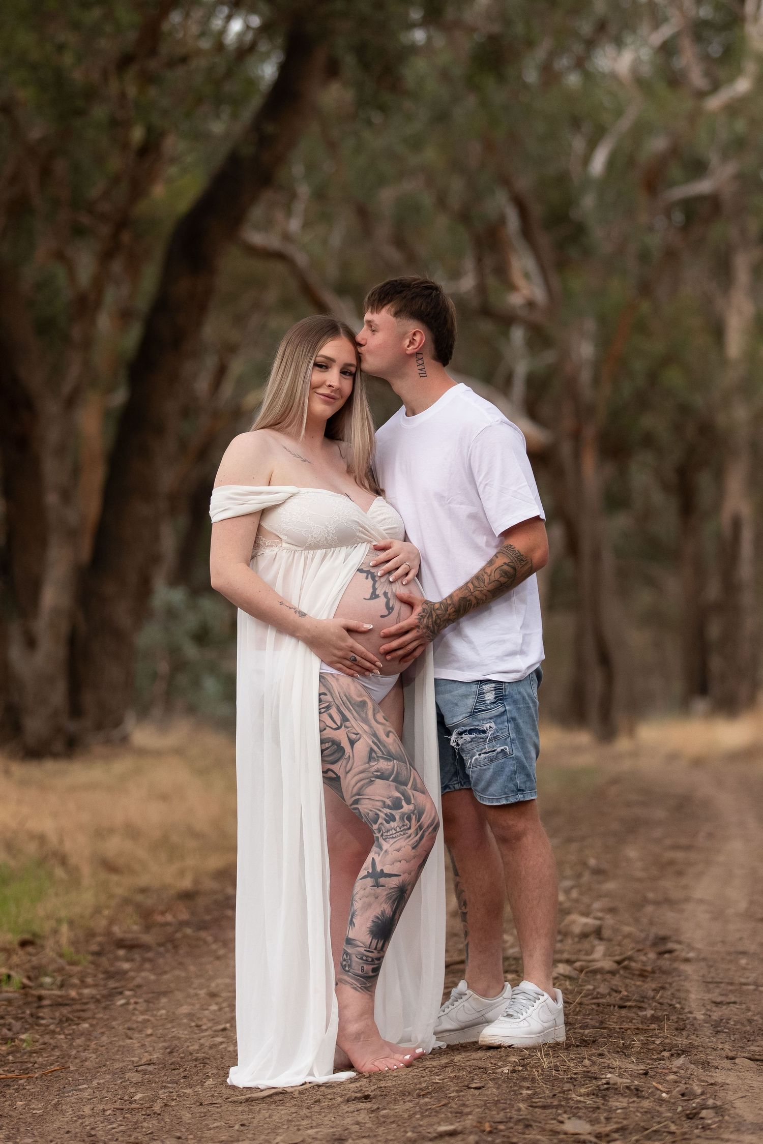 Tattooed couple in forest, pregnant woman in white dress, man kissing her cheek while holding baby bump.