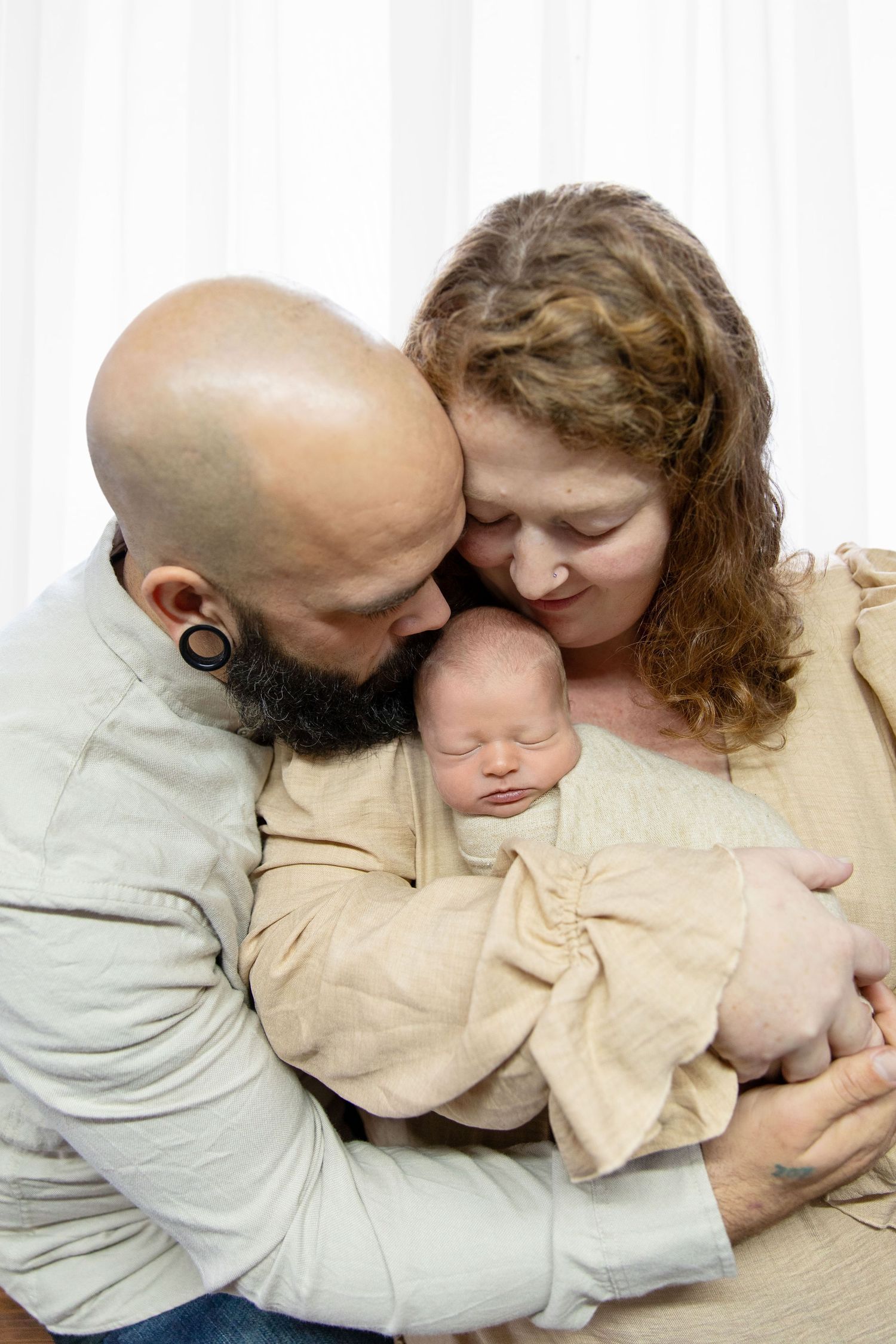 Bald bearded father and curly-haired mother tenderly kissing their sleeping newborn baby wrapped in beige cloth.