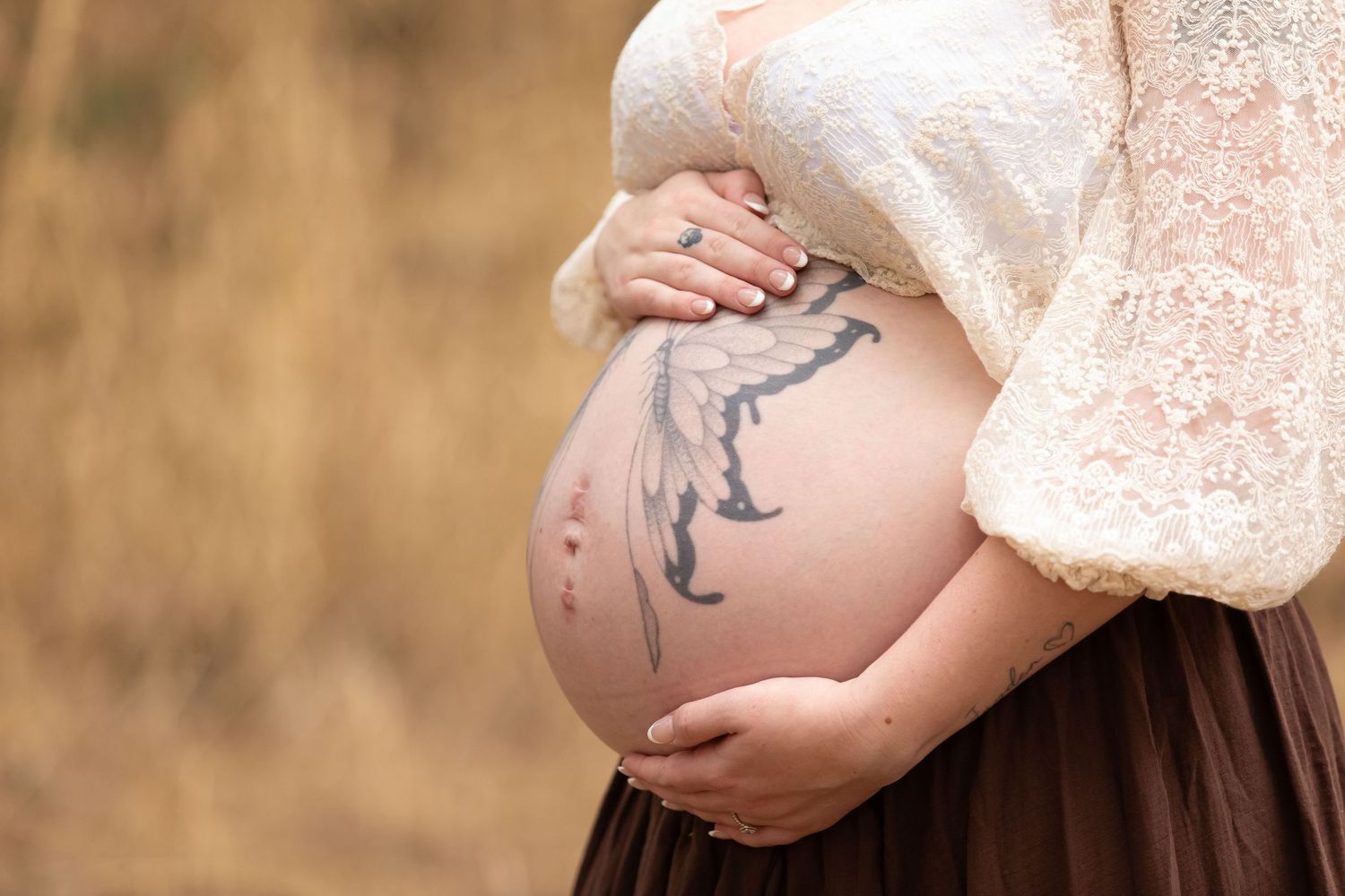Pregnant woman in lace top cradling tattooed baby bump outdoors in golden field.