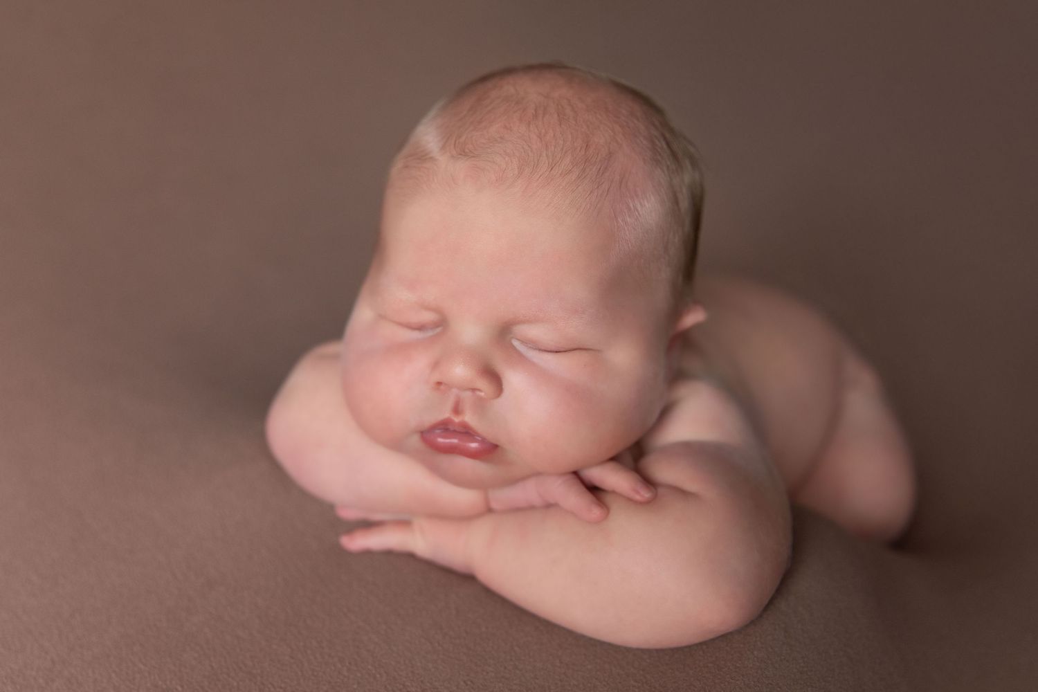 Peaceful newborn baby sleeping in froggy pose on brown backdrop, professional newborn photography studio portrait.