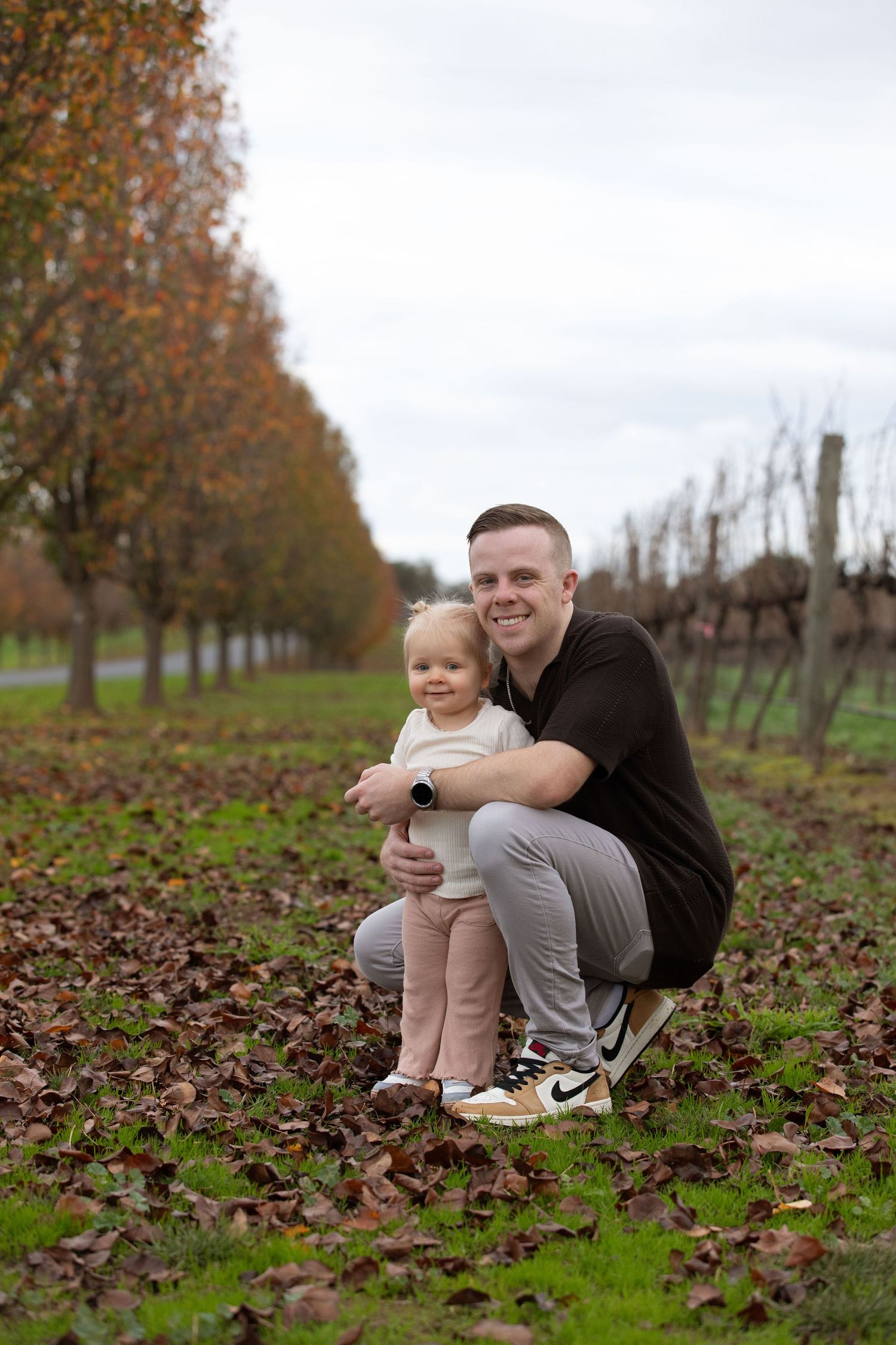 Man crouching with toddler girl among autumn leaves in a vineyard, smiling at camera on a cloudy day.
