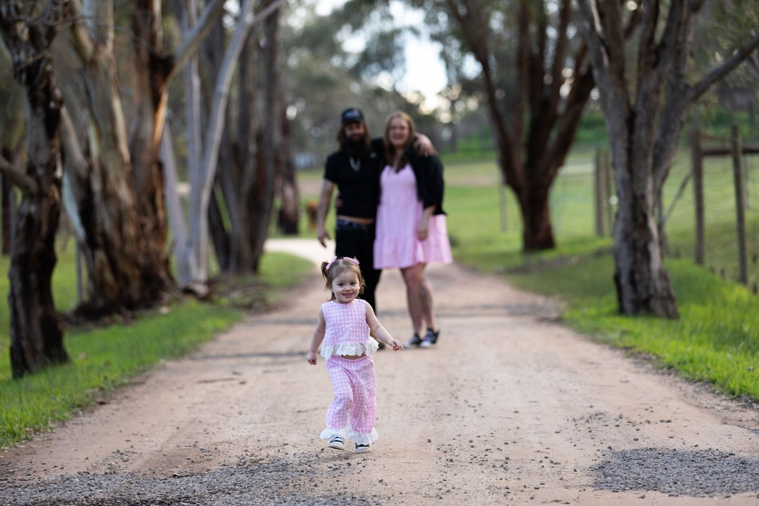 A young toddler girl in pink walks down a tree-lined dirt path as her parents smile in the background.