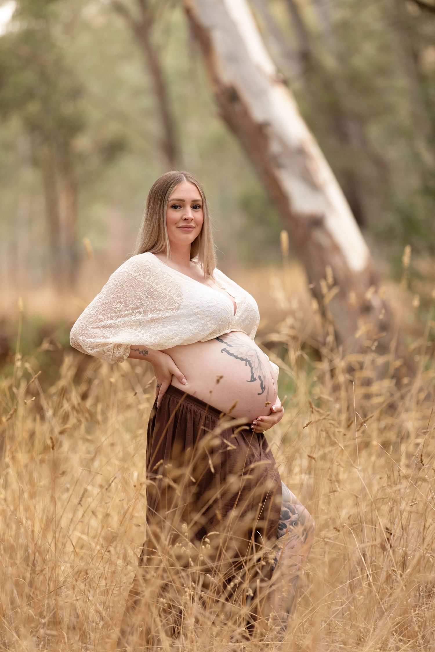 Pregnant woman in white wrap top and brown skirt holding baby bump in golden grass field with trees.