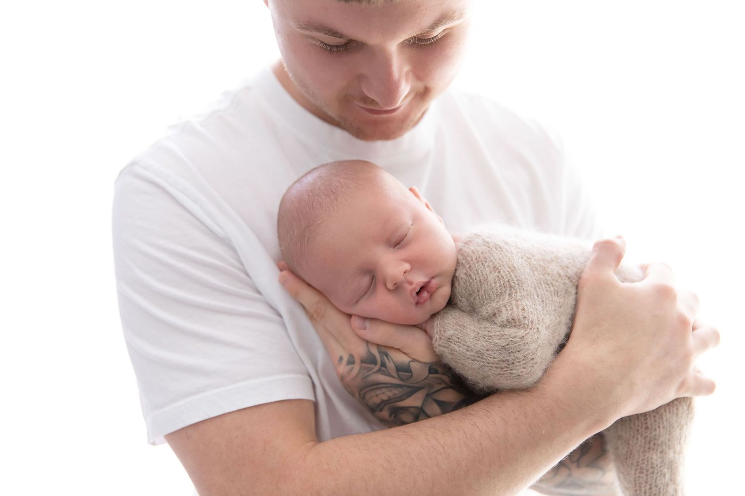 Father with tattoos tenderly holding sleeping newborn baby wrapped in beige knit blanket against white background.
