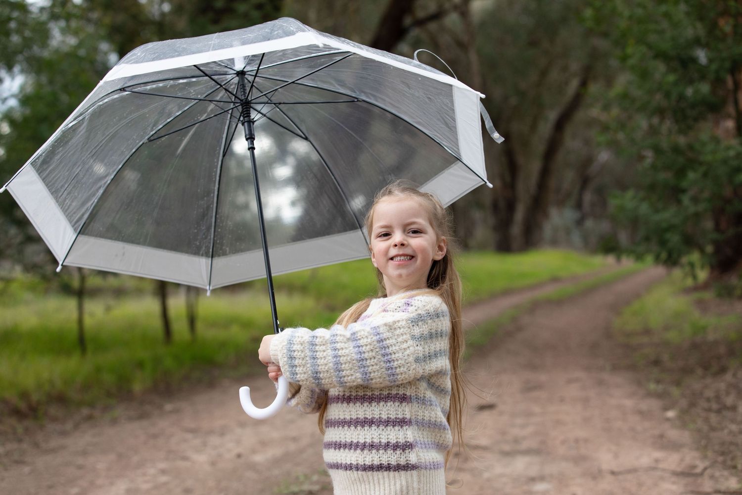 Smiling young girl holding a clear transparent umbrella on a forest path wearing a cosy striped sweater.