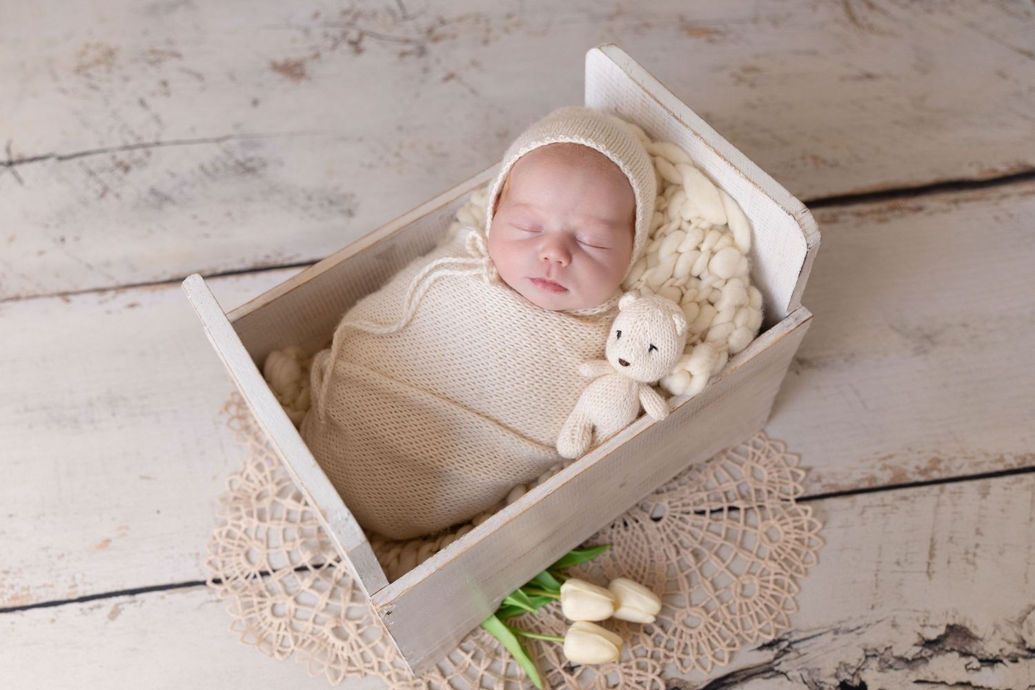 A sleeping newborn wrapped in beige swaddle lies in a white wooden box with a small stuffed animal and delicate lace accents.