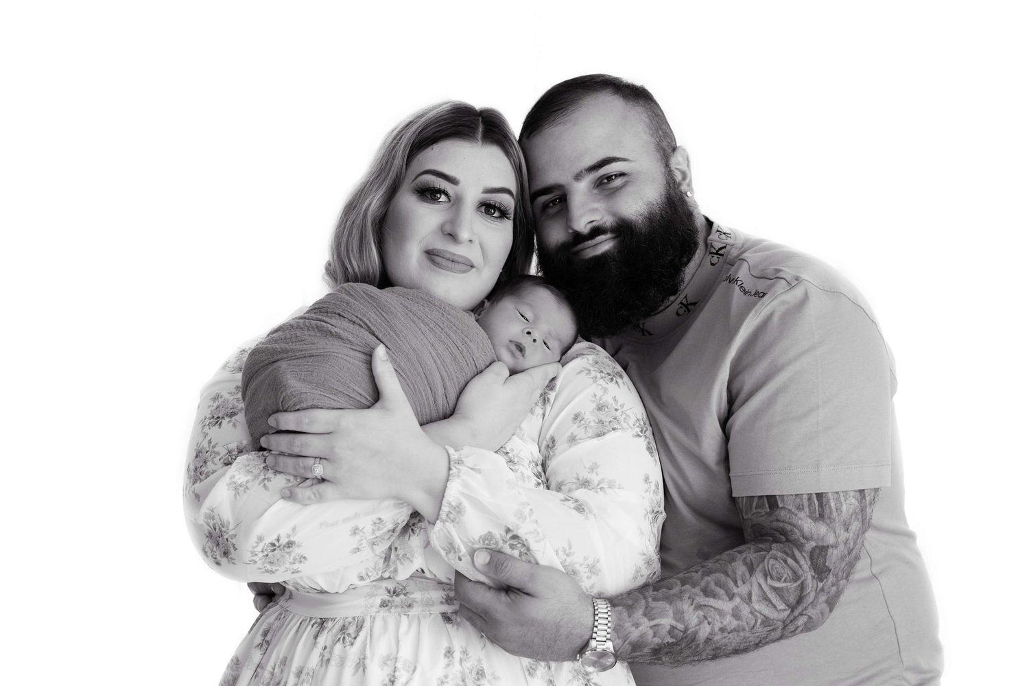 A black and white portrait shows a couple embracing their newborn, featuring a bearded man with tattoos and someone in a floral dress.