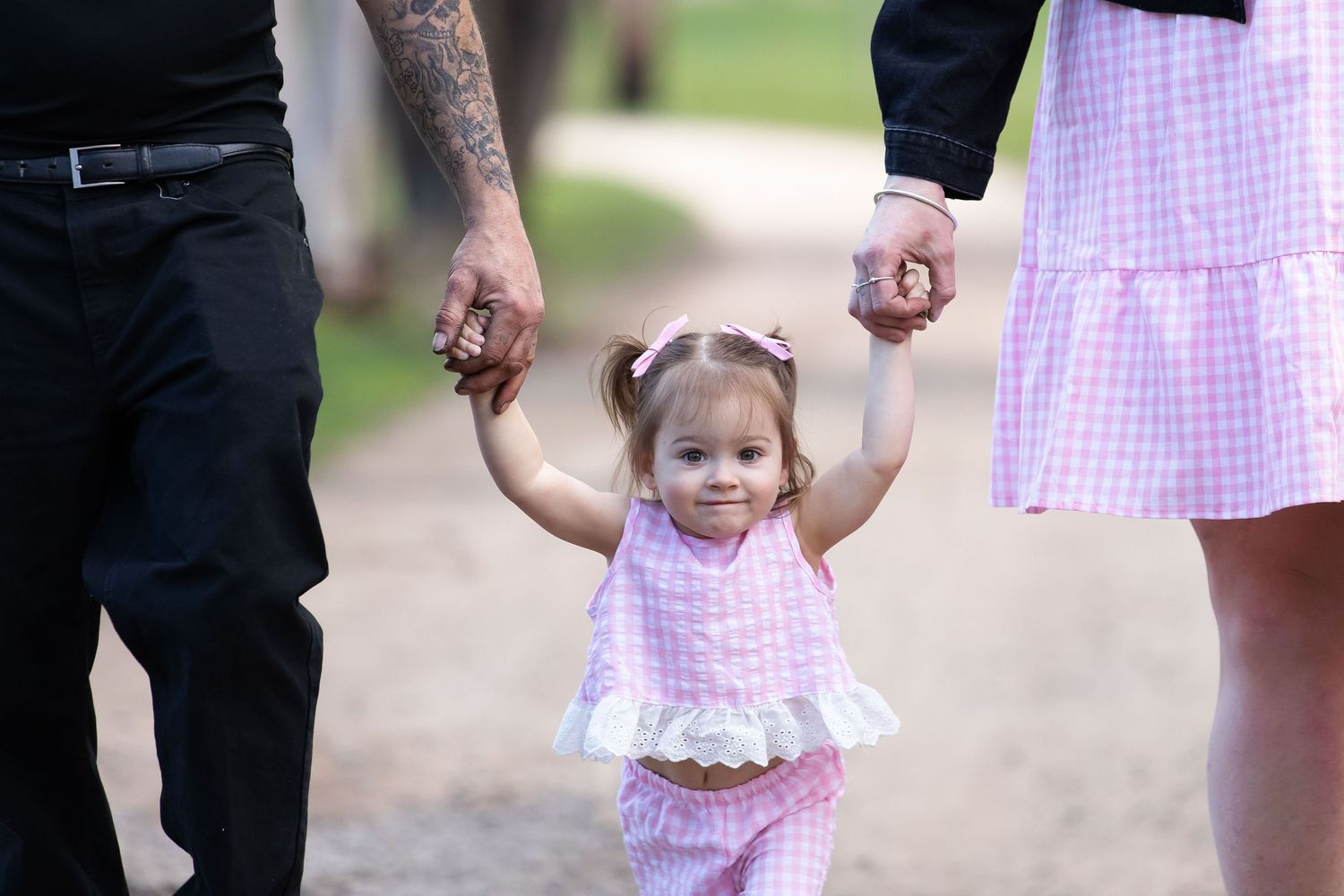 Toddler girl in pink outfit holds hands with parents while walking on a path outdoors.