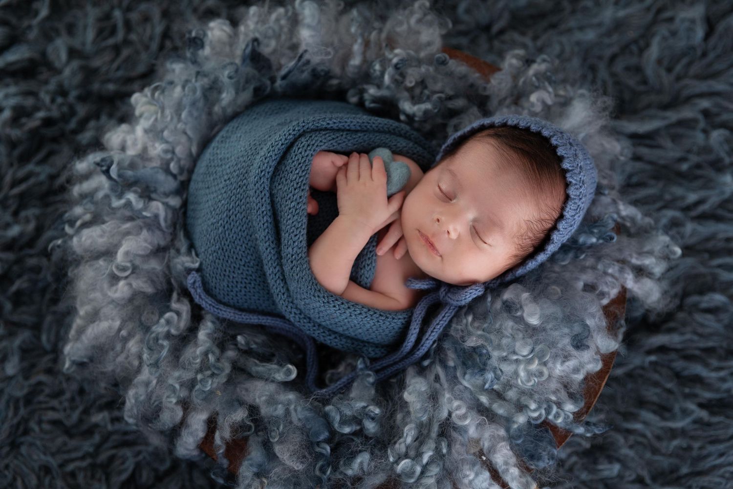 Sleeping newborn baby wrapped in soft blue swaddle blanket nestled on blue textured backdrop.