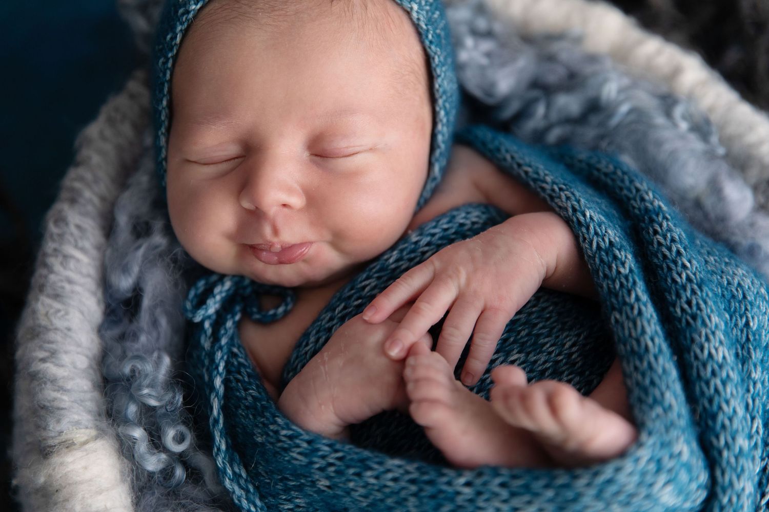 Sleeping newborn baby wrapped in a teal blue knit blanket and bonnet, peacefully resting in a cosy nest.