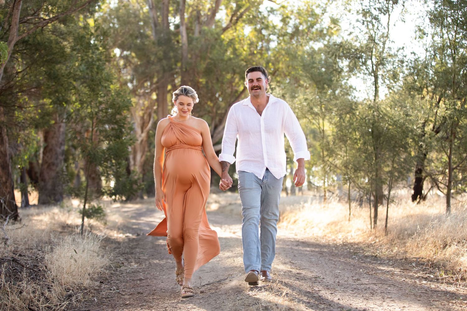 Pregnant woman in orange dress walks hand-in-hand with man in white shirt on a sunlit dirt path surrounded by trees.
