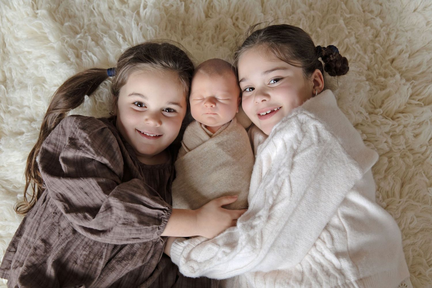 Two smiling girls lying on a fluffy rug with a swaddled newborn baby between them.