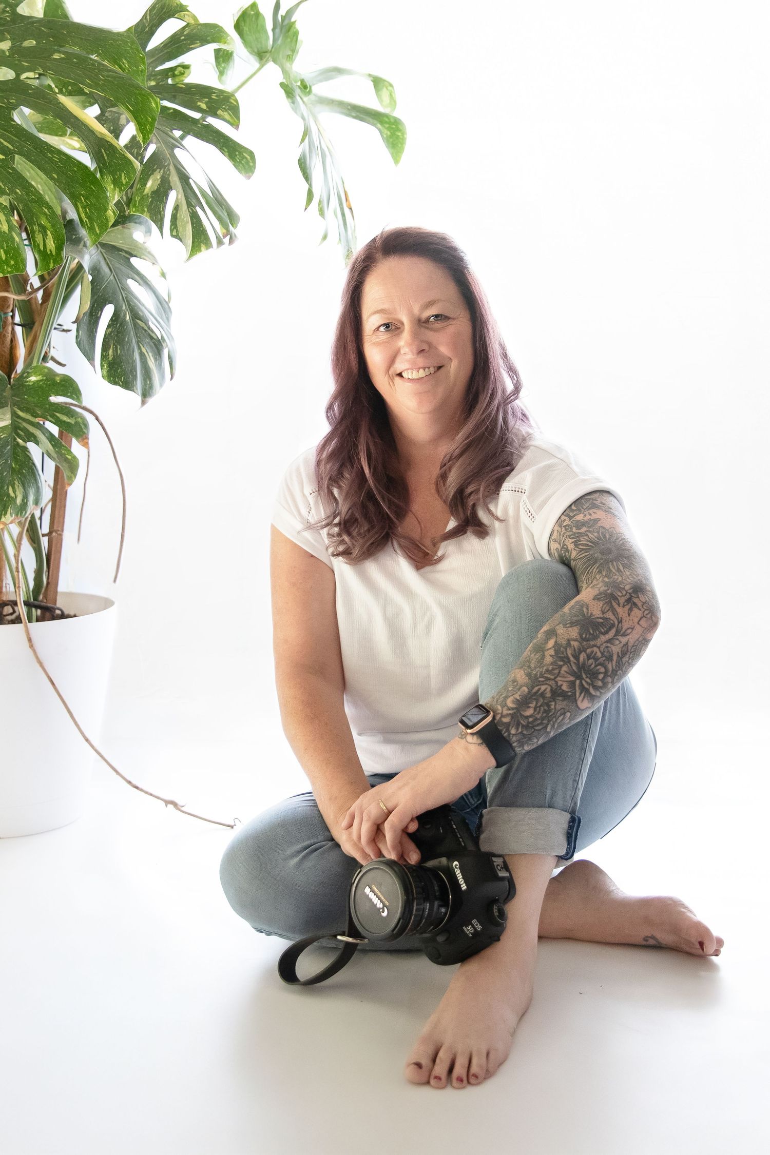 Woman with tattoo sleeve sits cross-legged holding DSLR camera, smiling, wearing white tee and jeans near monstera plant.
